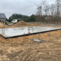 Wylany betonowy fundament z wystającymi prętami zbrojeniowymi, zabezpieczony czarną folią kubełkową, na działce z piaskiem i widocznym domem w tle.