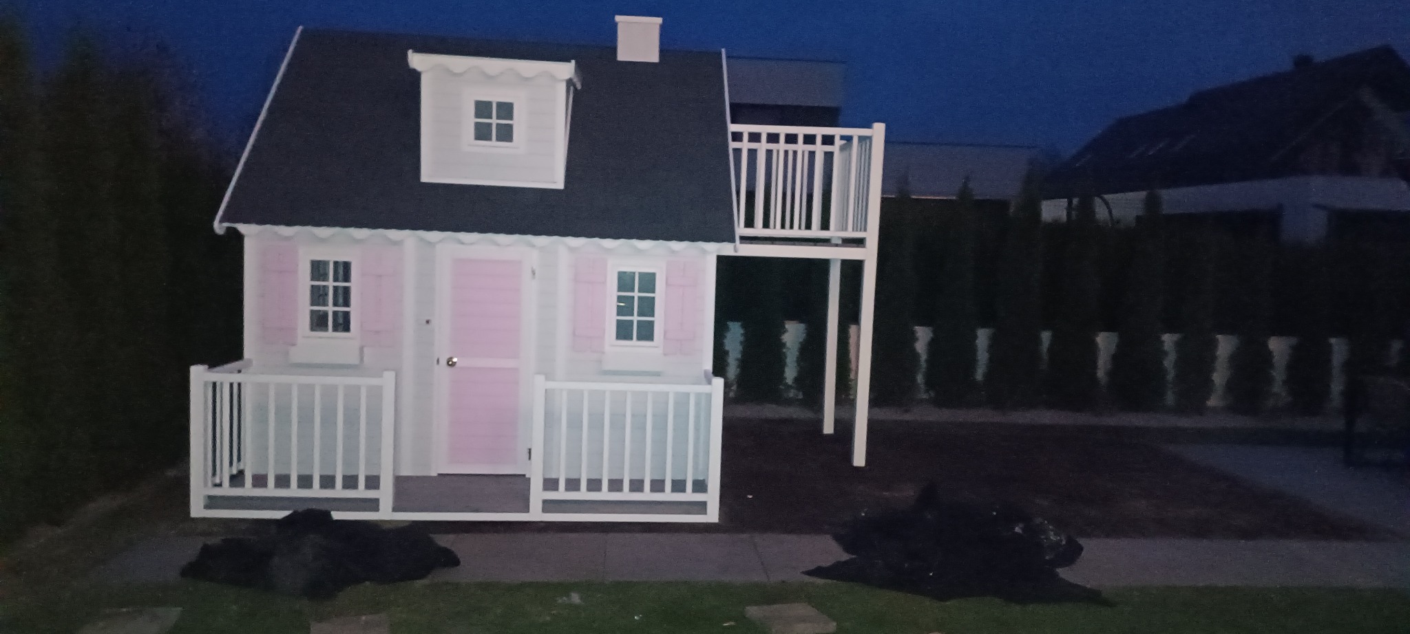 Biało-różowy domek dla dzieci z balkonem, ustawiony na trawniku, otoczony tujami, sfotografowany w słabym oświetleniu wieczornym. Widoczny fragment kostki brukowej.