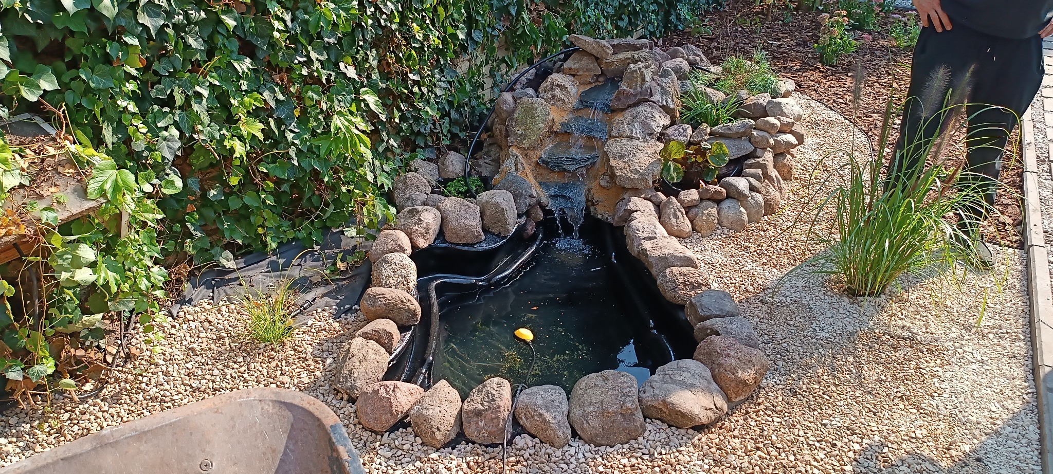 Oczko wodne w ogrodzie z kaskadą wodną, obłożone kamieniami i żwirem, otoczone zielenią. Woda w oczku jest czarna.