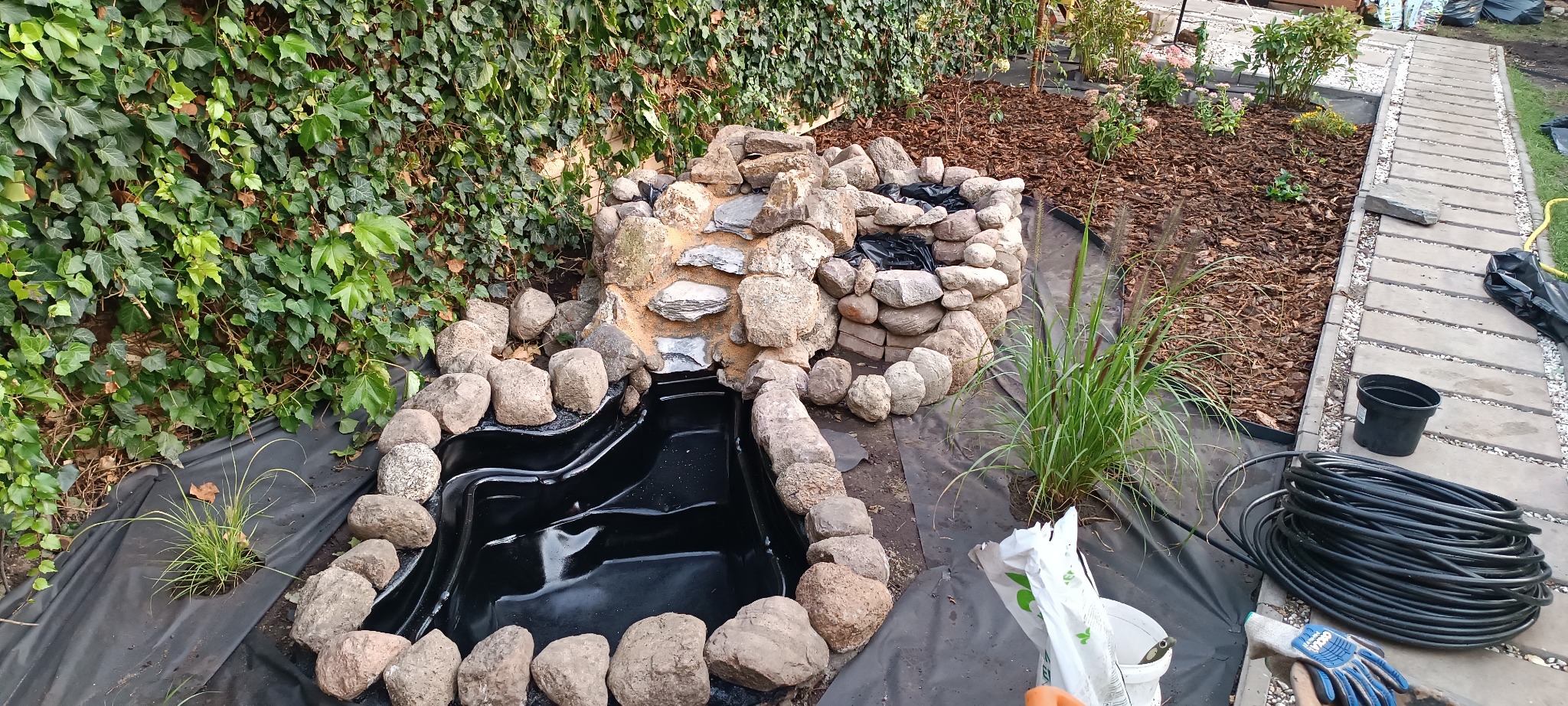 Budowa oczka wodnego z kaskadą: czarna folia, kamienie rzeczne, rośliny ozdobne, w tle ściana zieleni z bluszczu. Elementy systemu nawadniania obok.