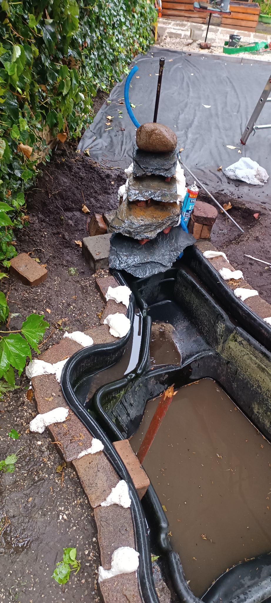 Budowa oczka wodnego z kaskadą w ogrodzie: czarne formy, kamienna kaskada, ceglana obudowa z pianką montażową. Etap prac przed uruchomieniem.