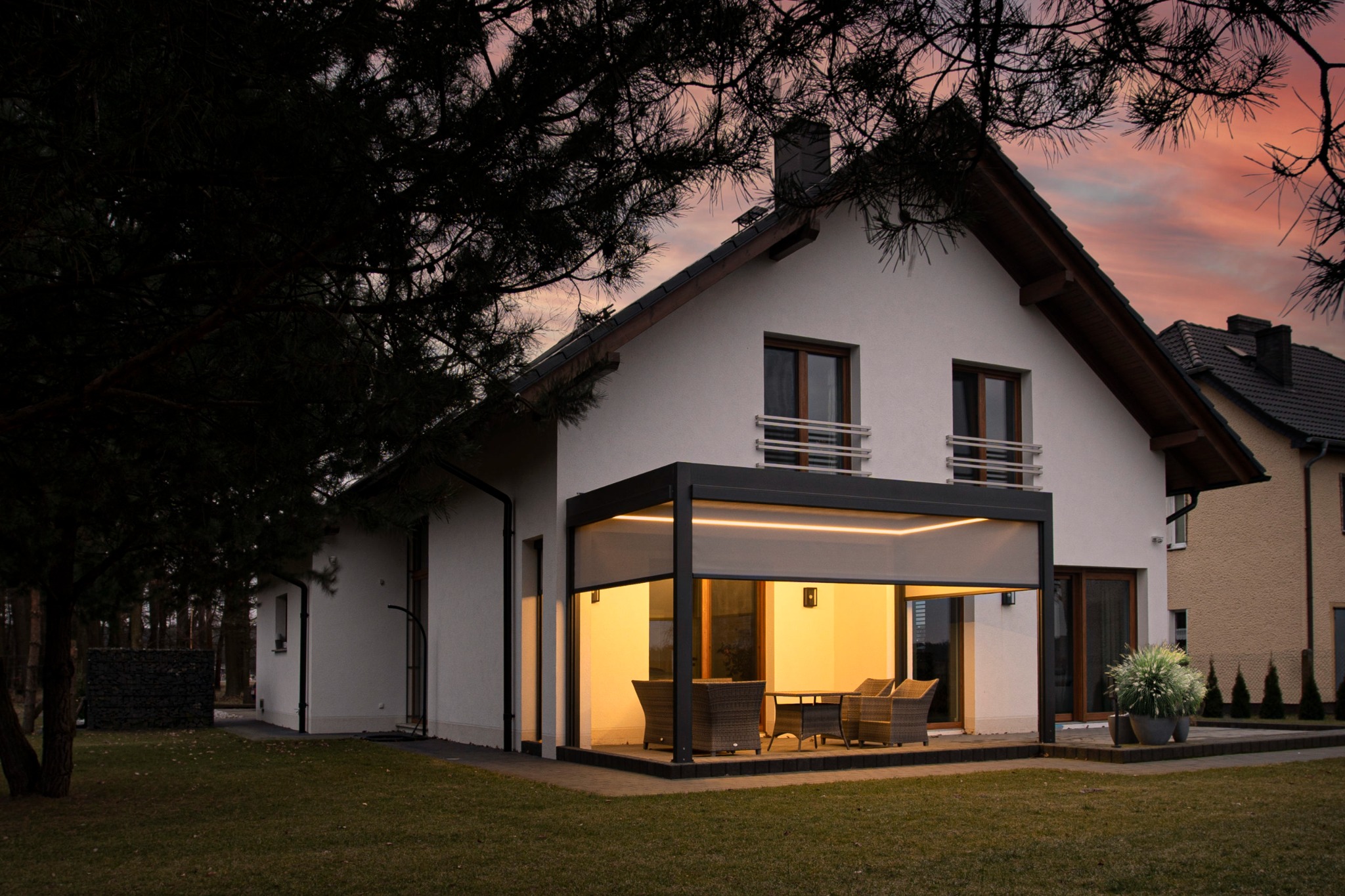 Nowoczesna pergola z metalową konstrukcją przylegająca do białego domu jednorodzinnego, oświetlona ciepłym światłem, z widocznymi meblami ogrodowymi i fragmentem zachodzącego słońca na niebie.