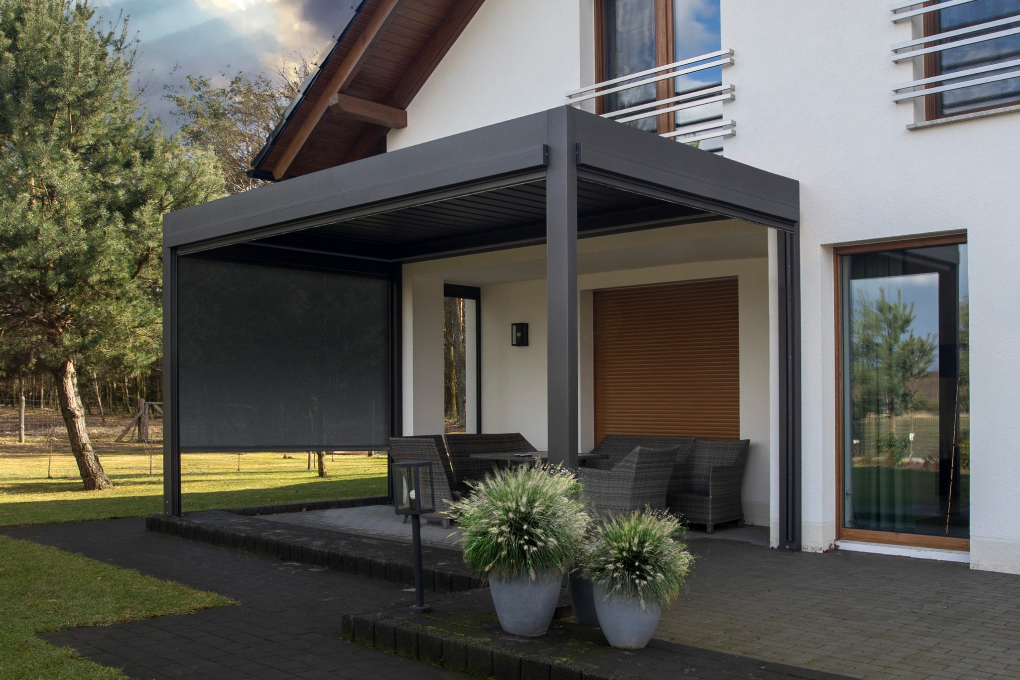 Nowoczesna, antracytowa pergola metalowa z roletą przeciwsłoneczną, przylegająca do białego domu z dużymi oknami, na pierwszym planie dwa ozdobne krzewy w szarych donicach.