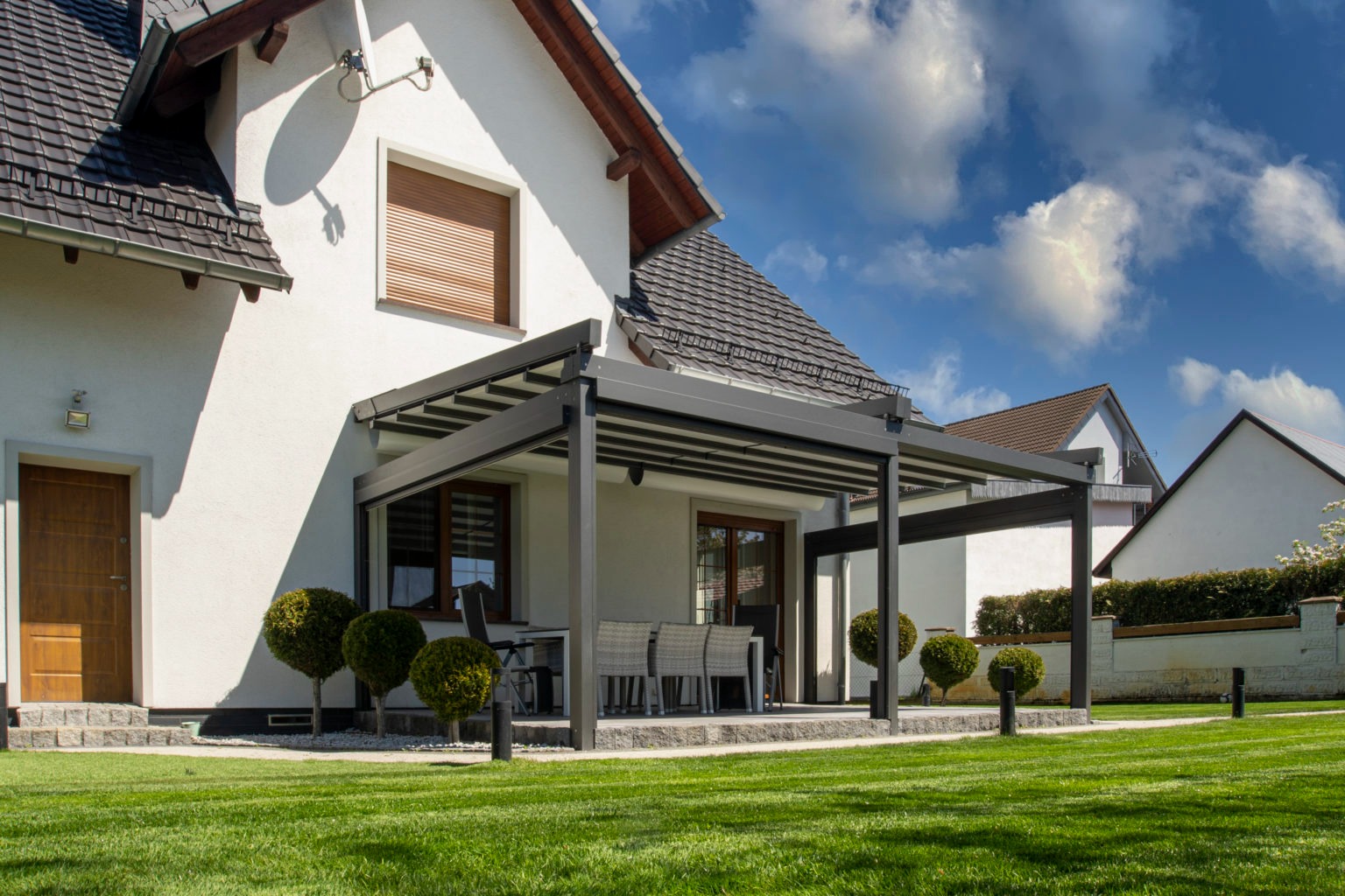 Nowoczesna, grafitowa pergola z metalu przylegająca do białego domu z zadbanym trawnikiem i kulistymi krzewami.