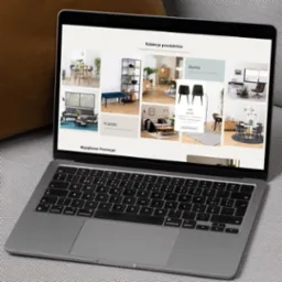 Laptop wyświetlający kolekcję produktów meblowych, w tym biurka i krzesła, na tle sofy z poduszką.
