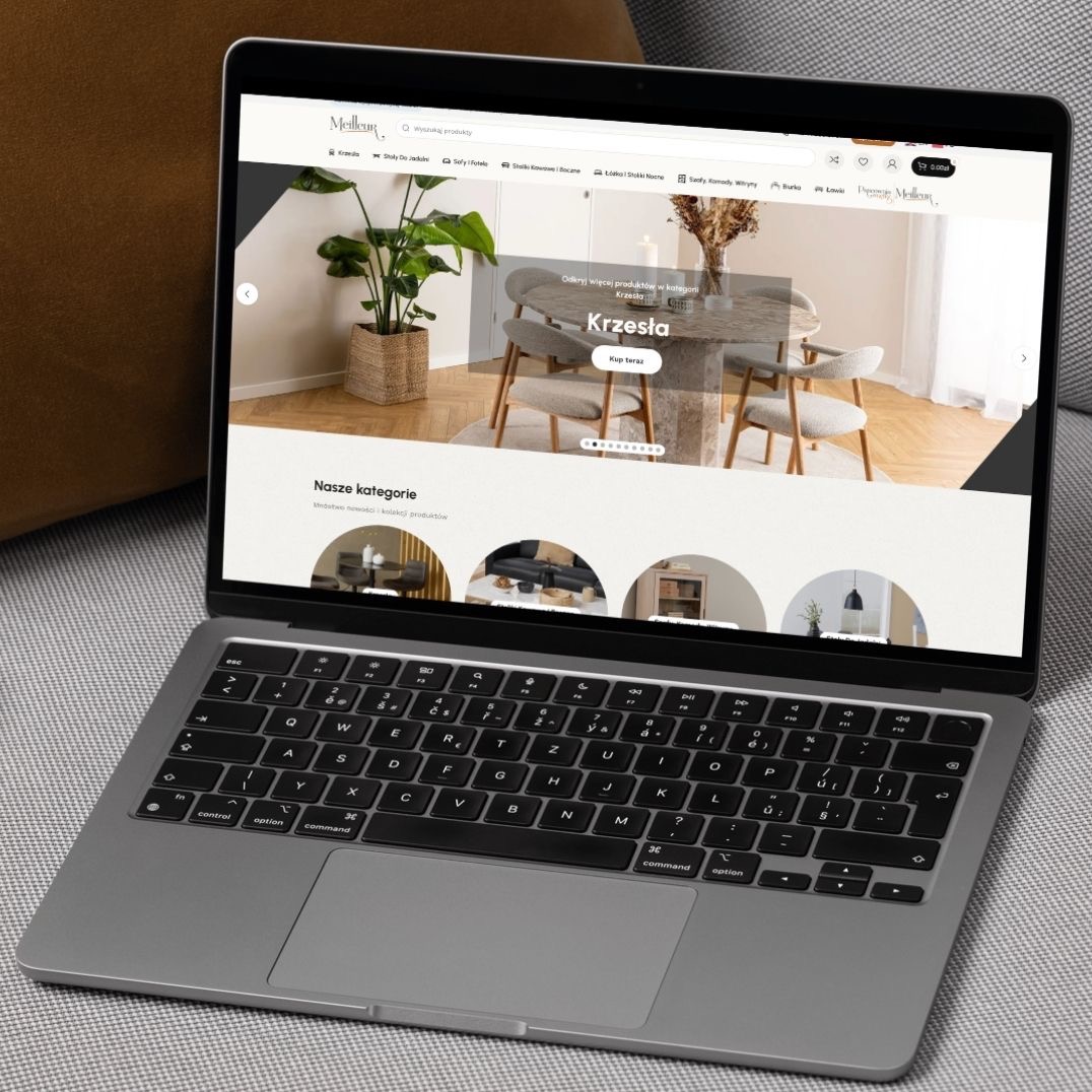 Laptop wyświetla stronę internetową Meilleur.pl z aranżacją wnętrza: okrągły stół z krzesłami, roślina doniczkowa i minimalistyczny design. Widoczny fragment klawiatury laptopa i szare tło kanapy.