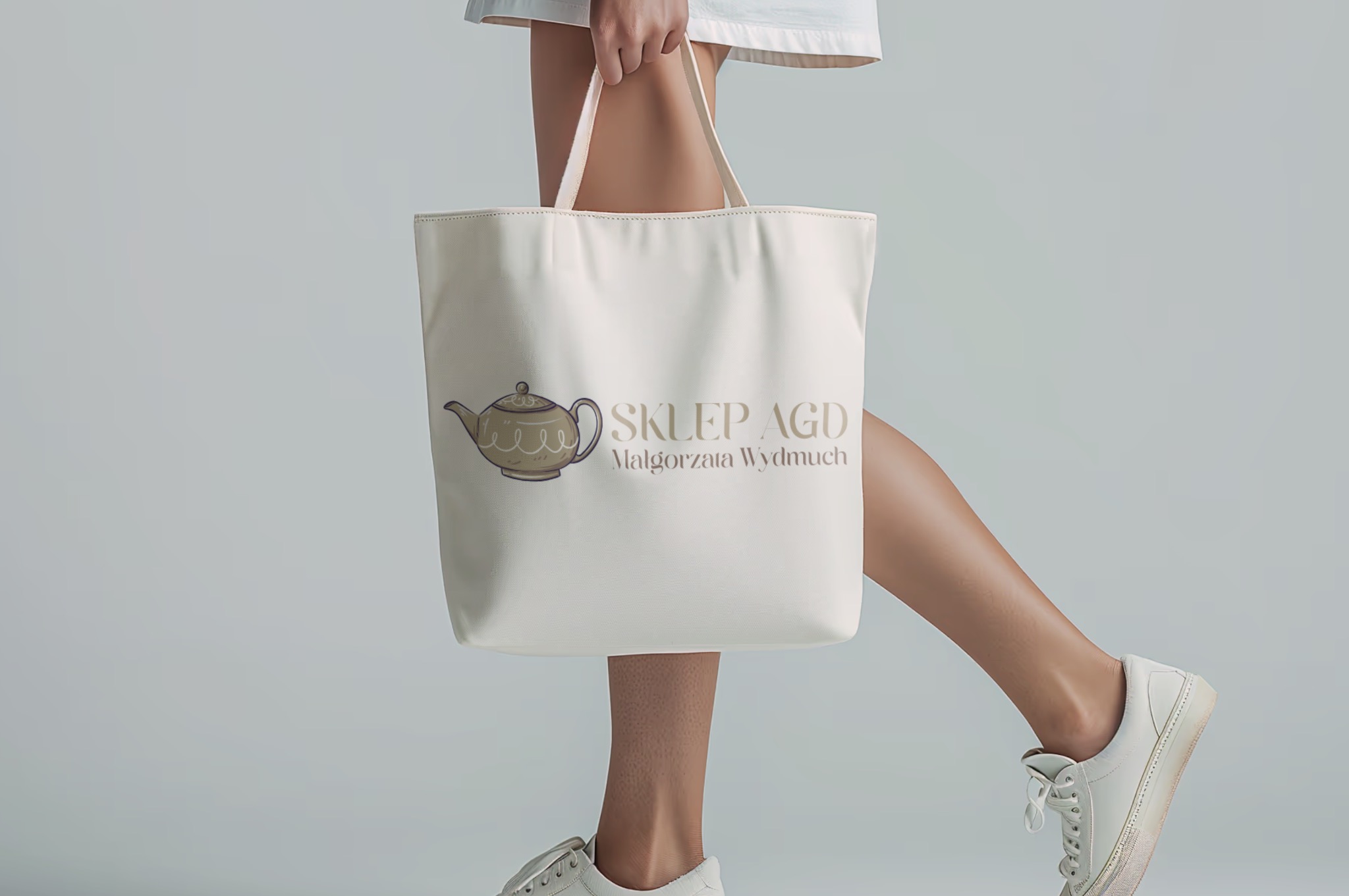 Beżowa torba shopper z nadrukiem w postaci czajnika i napisu 'Sklep AGD Małgorzata Wydmuch', trzymana przez modelkę w białej sukience i sneakersach na szarym tle.