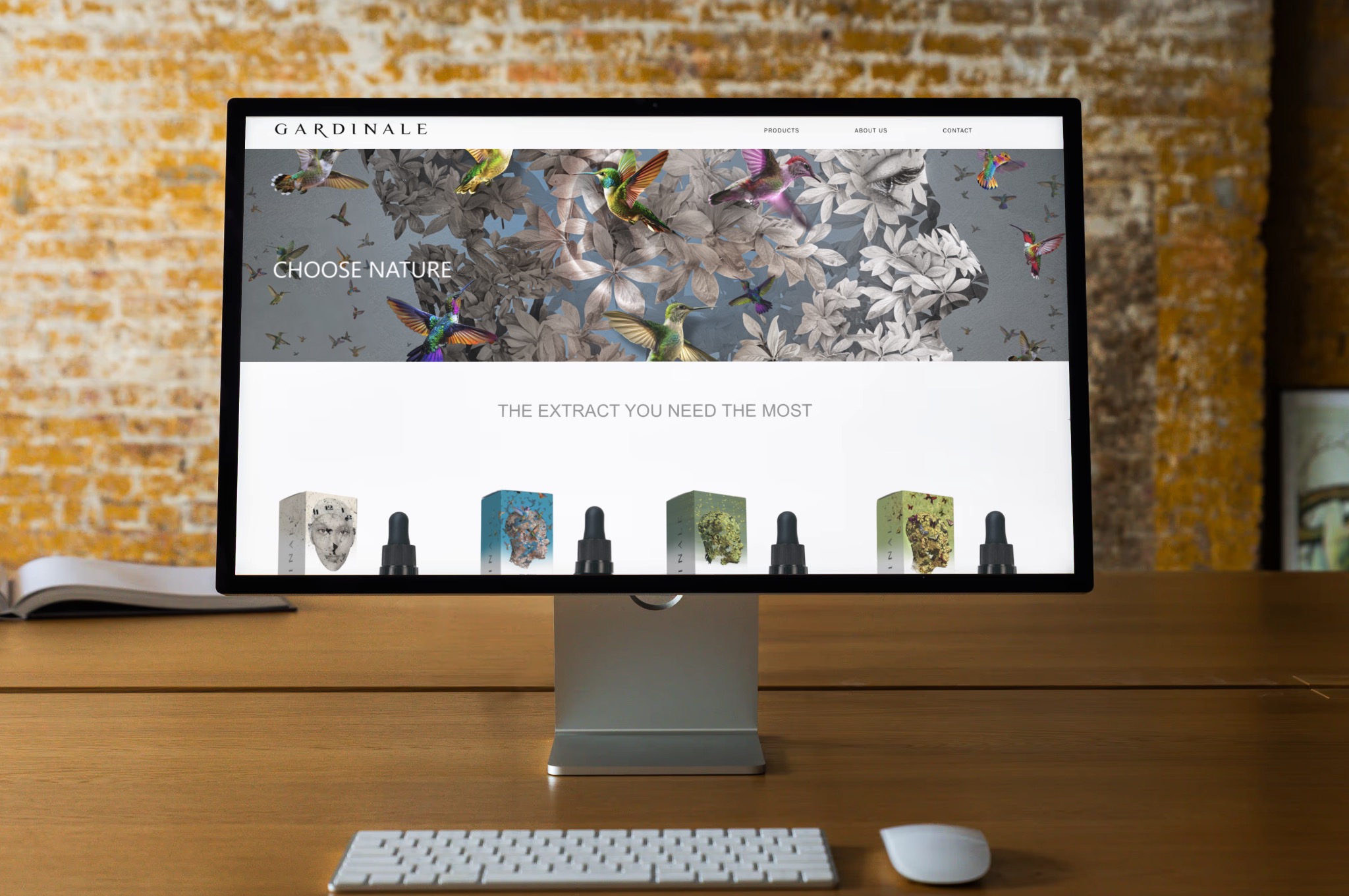 Monitor wyświetla stronę internetową z motywem natury, kolibry i kwiaty w tle nagłówka, poniżej prezentacja produktów w pudełkach, na drewnianym biurku z klawiaturą i myszką.