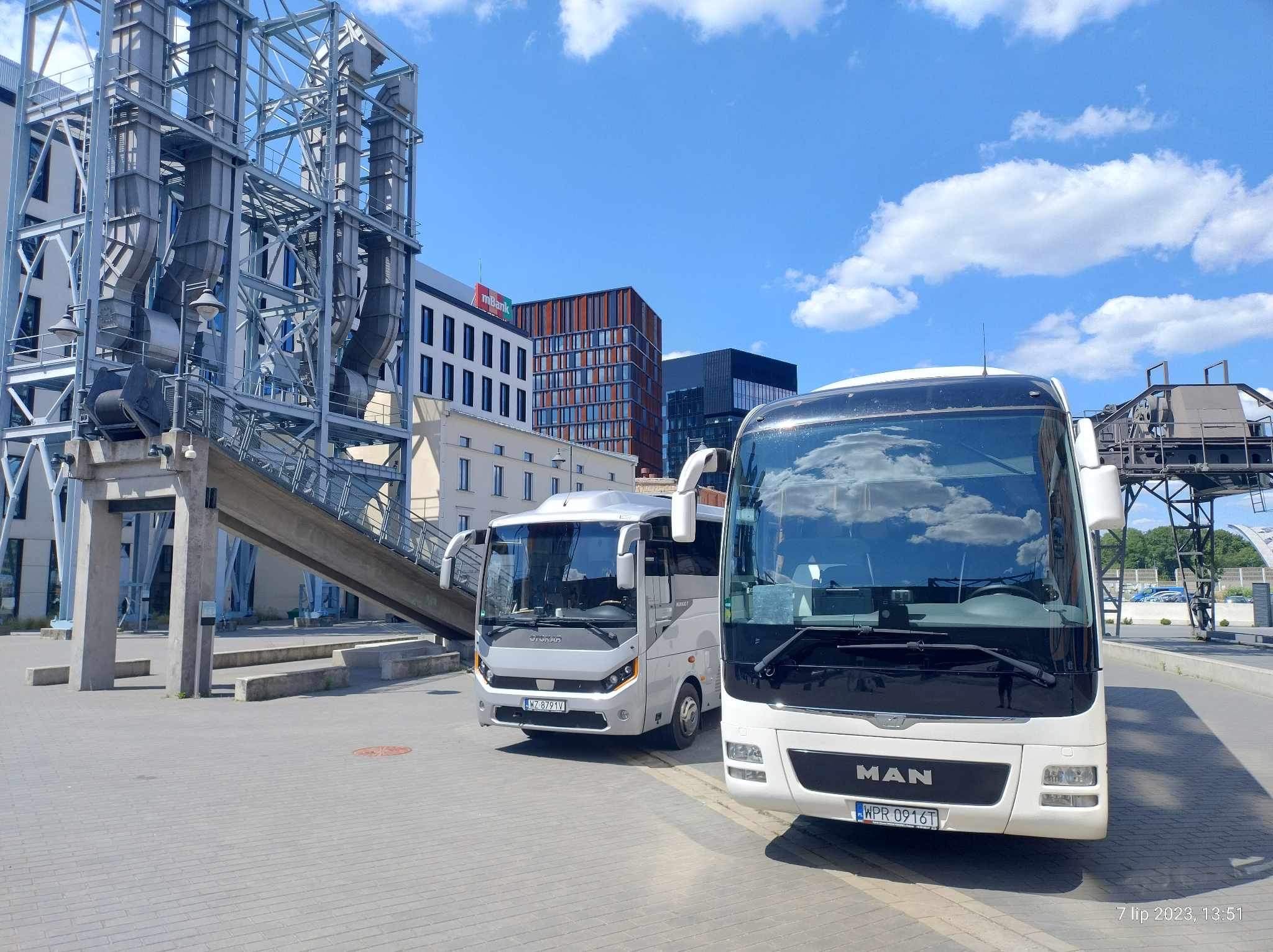 Dwa białe autokary MAN na tle industrialnego krajobrazu z metalową konstrukcją i nowoczesnymi budynkami w słoneczny dzień.