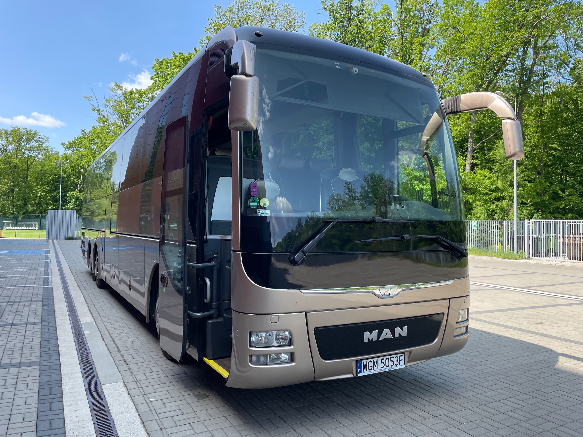 Nowoczesny autokar MAN w kolorze szampańskim, zaparkowany na kostce brukowej z otwartymi drzwiami wejściowymi, widoczne tablice rejestracyjne WGM 5053F.
