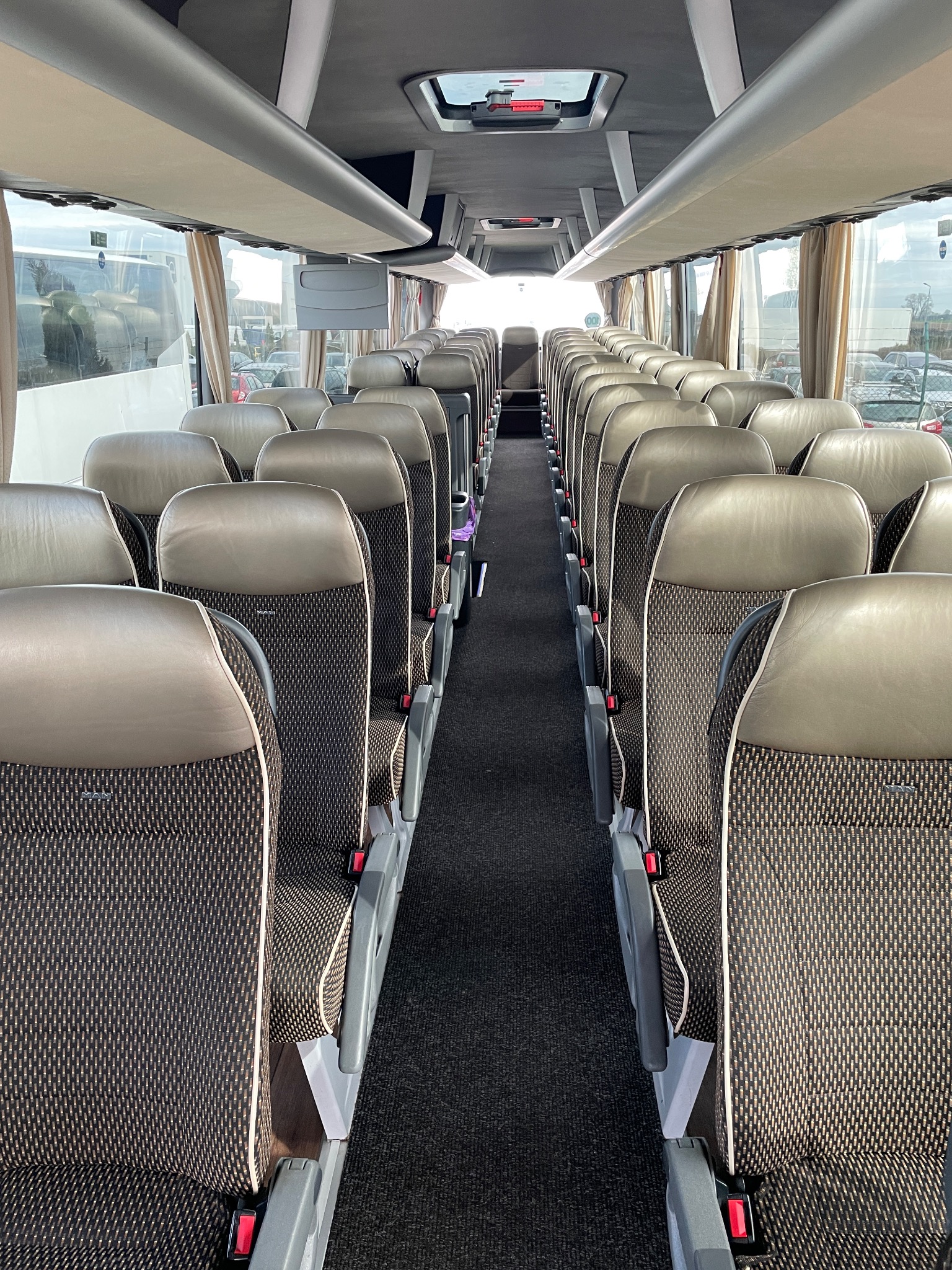 Wnętrze autobusu z rzędami brązowo-czarnych foteli z zagłówkami, widok na korytarz w kierunku jasnego światła na końcu.