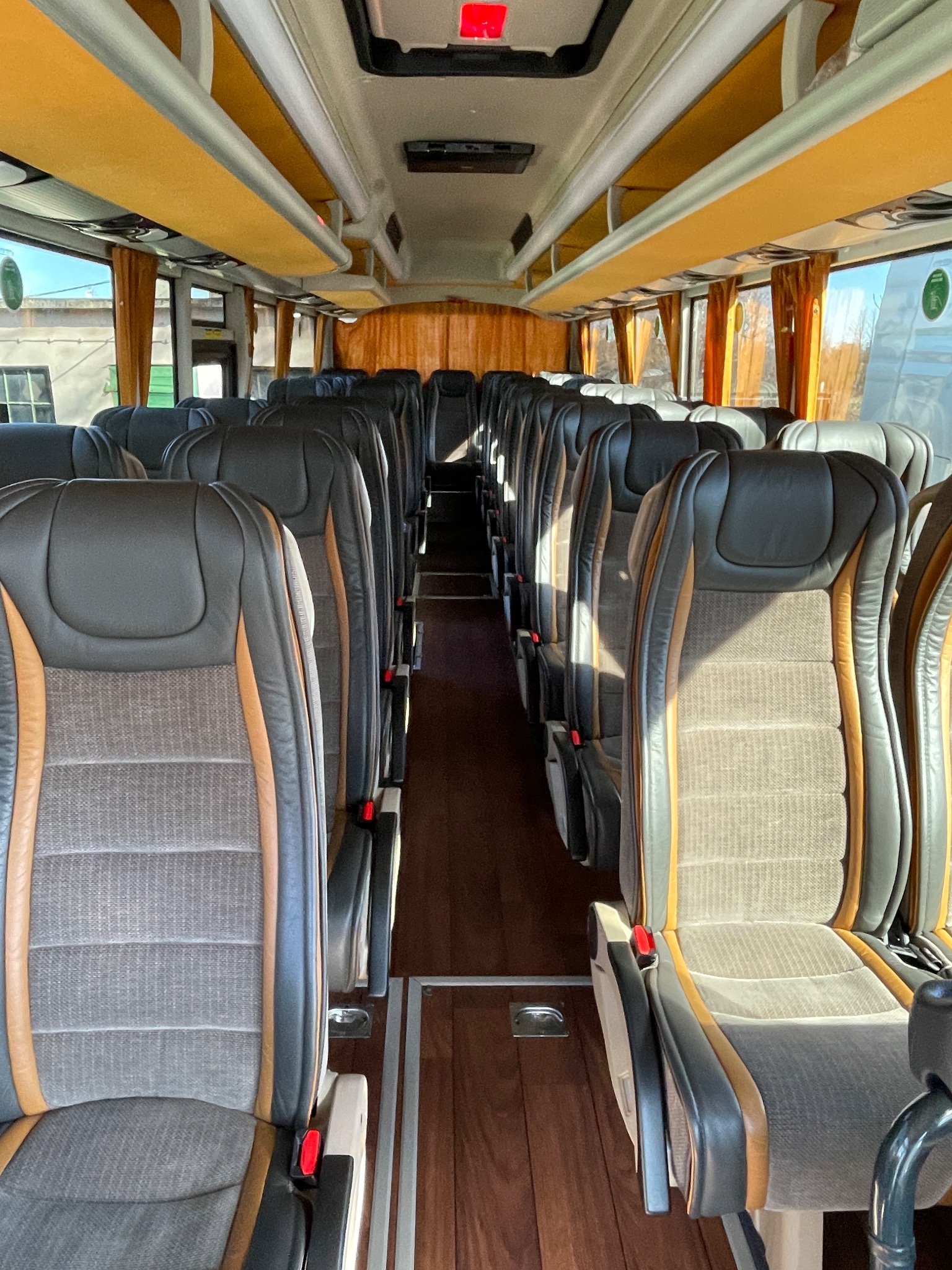 Wnętrze autobusu turystycznego z rzędami szarych foteli z brązowymi akcentami, drewnianą podłogą i żółtymi zasłonami w oknach.