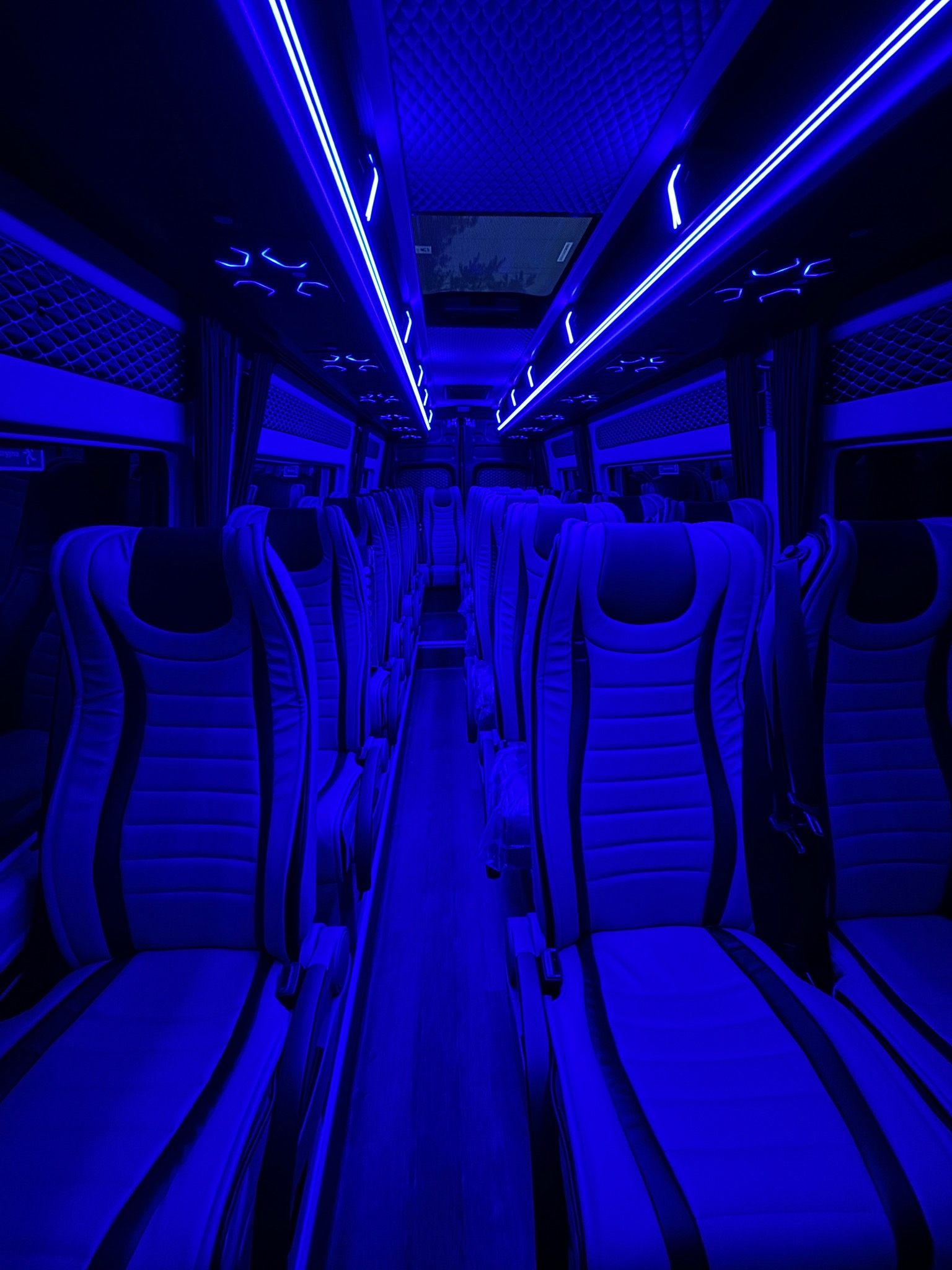 Wnętrze luksusowego busa oświetlone niebieskim światłem LED; widoczne rzędy skórzanych foteli z czarnymi wstawkami, podświetlany sufit z pikowaną fakturą i okna z zasłonami.
