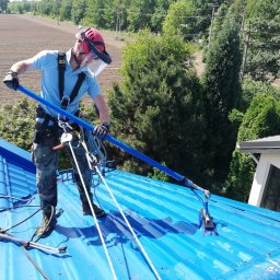FCSG BARTŁOMIEJ REKOS - Pracownik w uprzęży i kasku z osłoną twarzy czyści niebieski dach za pomocą szczotki na długim wysięgniku; w tle drzewa i pole.