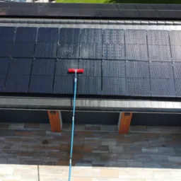 Widok z g&oacute;ry na panele słoneczne na dachu garażu podczas czyszczenia za pomocą szczotki na długim kiju, widoczne ślady wody i smug na panelach, na dachu r&oacute;wnież zamontowane dodatkowe panele.