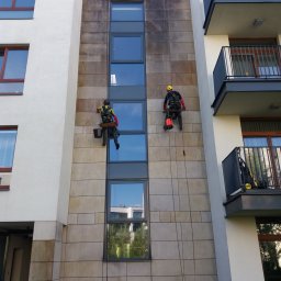FCSG BARTŁOMIEJ REKOS - Dwóch pracowników na linach czyści elewację budynku, widoczne różnice w czystości między umytą a brudną częścią ściany, zabezpieczenie folią na dole.