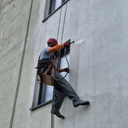 Pracownik w kasku i kombinezonie, zabezpieczony linami, myje elewację budynku pod ciśnieniem, widoczne ślady czyszczenia na jasnej fasadzie.