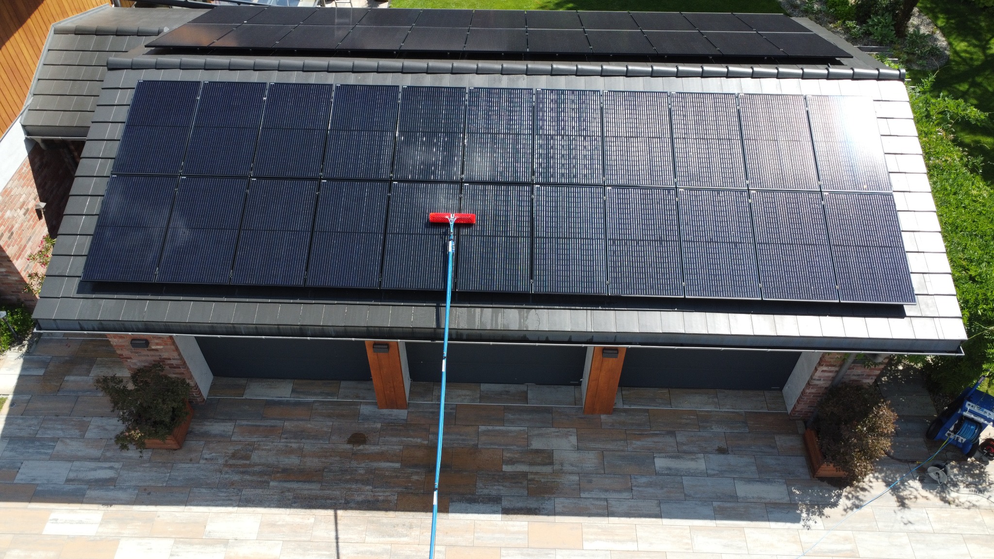 Widok z góry na panele słoneczne na dachu garażu podczas czyszczenia za pomocą szczotki na długim kiju, widoczne ślady wody i smug na panelach, na dachu również zamontowane dodatkowe panele.