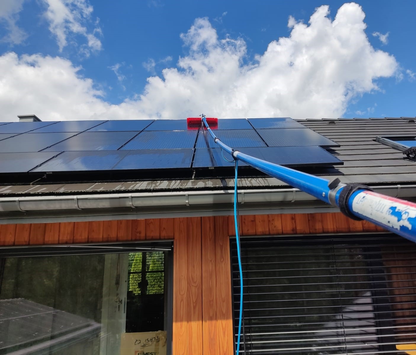 Czyszczenie paneli słonecznych na dachu domu za pomocą szczotki na teleskopowym kiju, błękitne niebo z chmurami w tle.