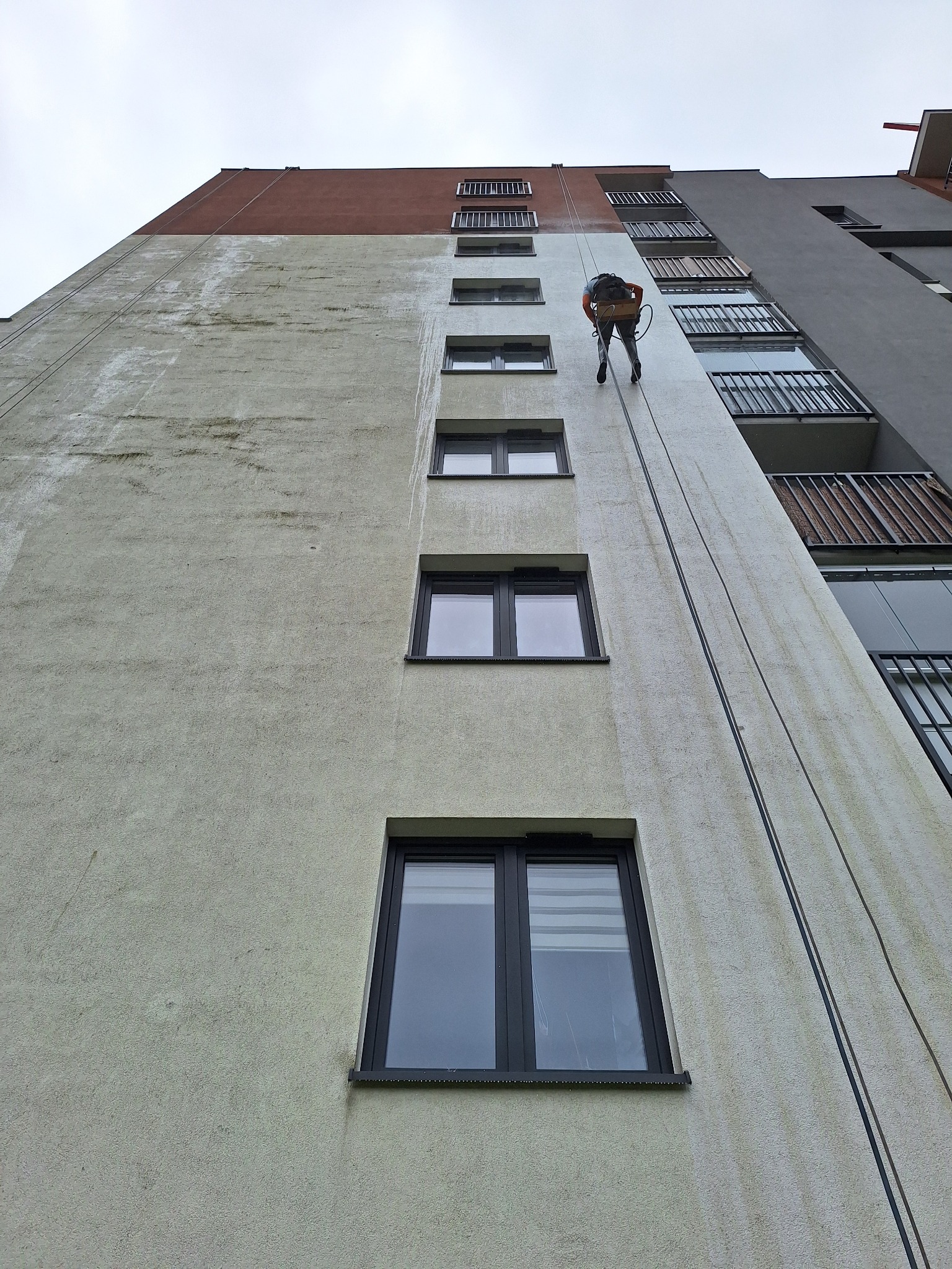 Pracownik na linach alpinistycznych czyści elewację wysokiego budynku, widoczne zacieki i brud na jasnej ścianie przed czyszczeniem, w tle szare balkony i fragment ceglanej ściany.