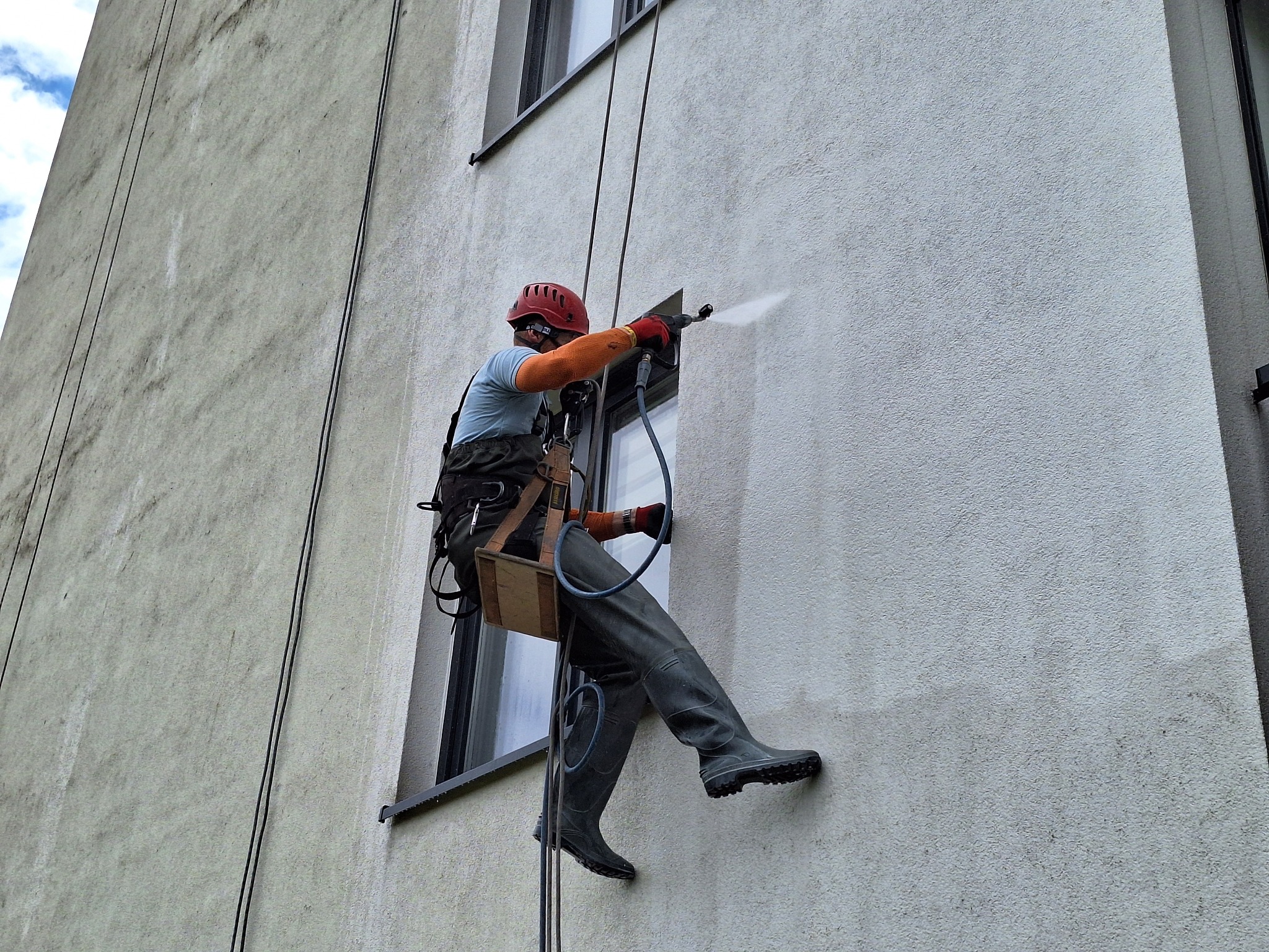 Pracownik w kasku i kombinezonie, zabezpieczony linami, myje elewację budynku pod ciśnieniem, widoczne ślady czyszczenia na jasnej fasadzie.