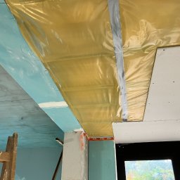 Paweł Jonczyk JON-TECH - Montaż sufitu podwieszanego: widoczne płyty gipsowo-kartonowe mocowane do folii paroizolacyjnej za pomocą wkrętarki akumulatorowej, fragment drabiny malarskiej w tle.