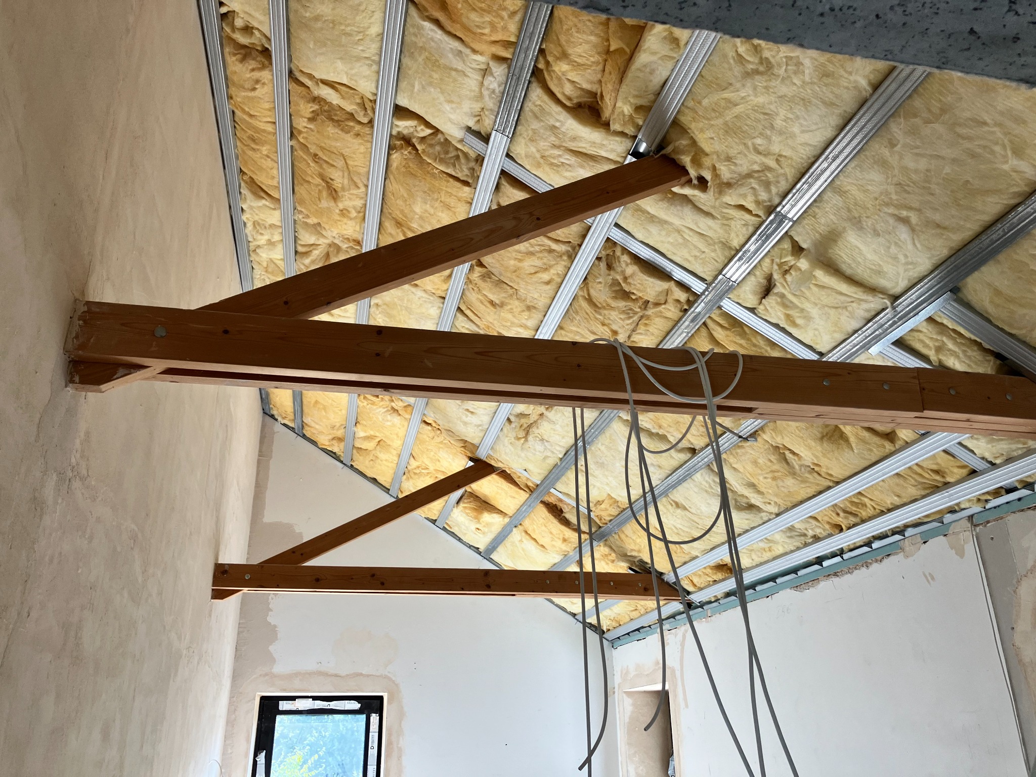 Montaż izolacji termicznej poddasza z widoczną konstrukcją drewnianą i metalowym stelażem, przewody elektryczne zwisają z sufitu.