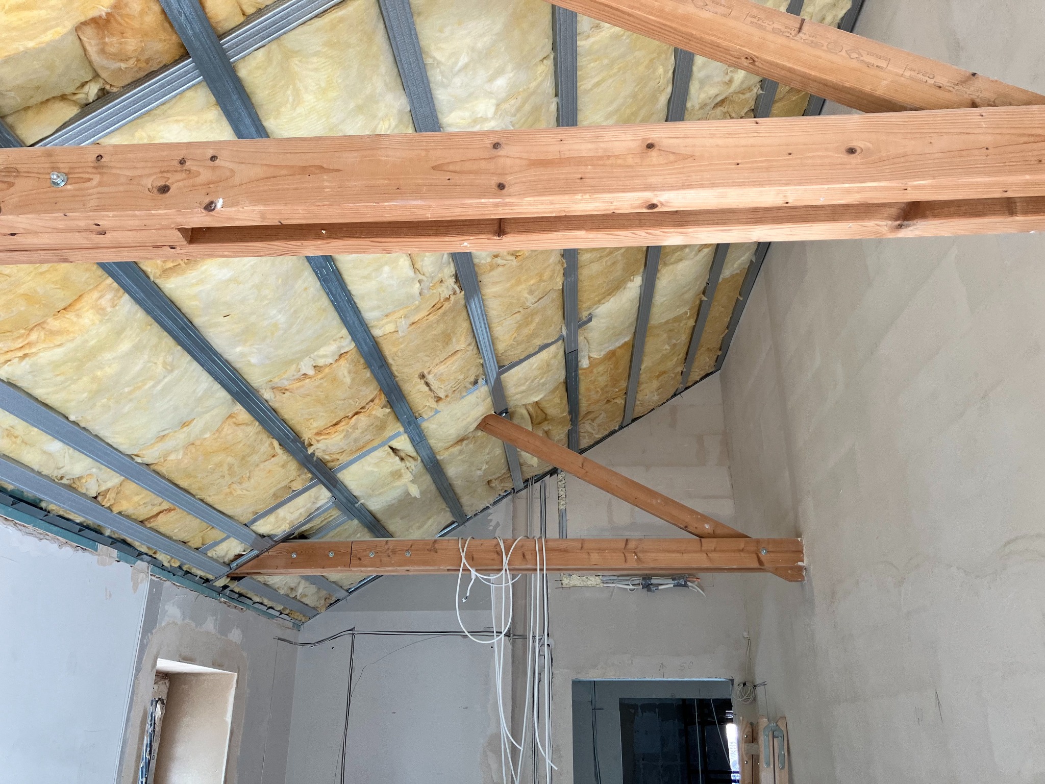 Wnętrze w trakcie remontu: izolacja z wełny mineralnej zamontowana między metalowymi profilami sufitowymi, drewniane belki wzmacniające, widoczne kable elektryczne i surowe ściany wymagające...