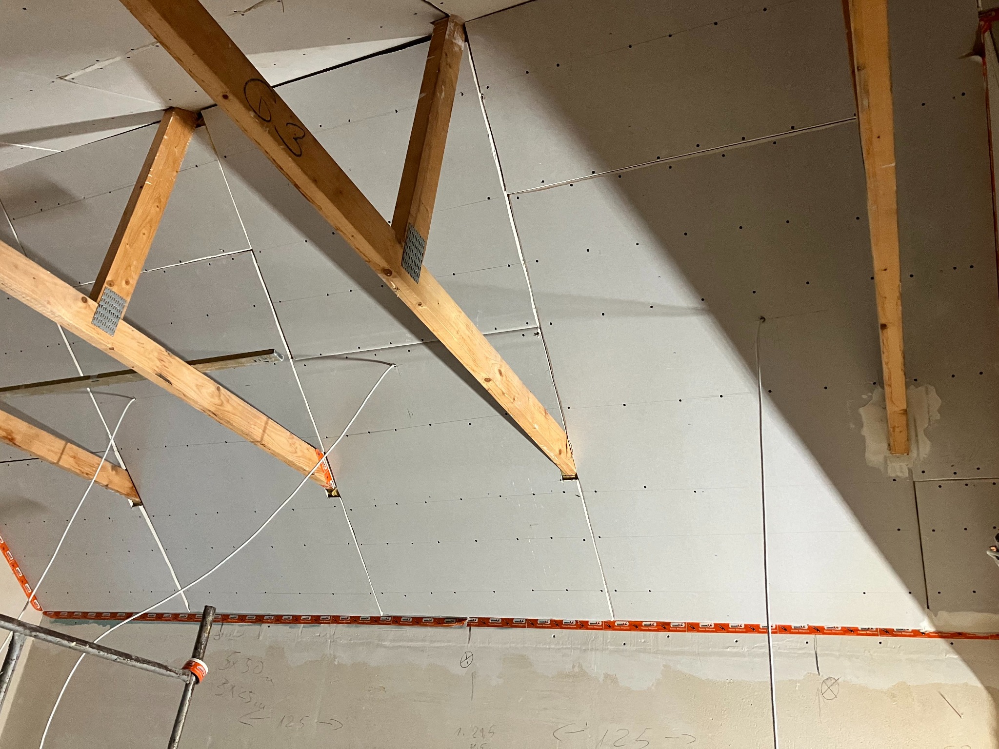 Montaż płyt gipsowo-kartonowych na poddaszu, widoczne drewniane krokwie, profile i instalacja elektryczna zabezpieczona taśmą ostrzegawczą.