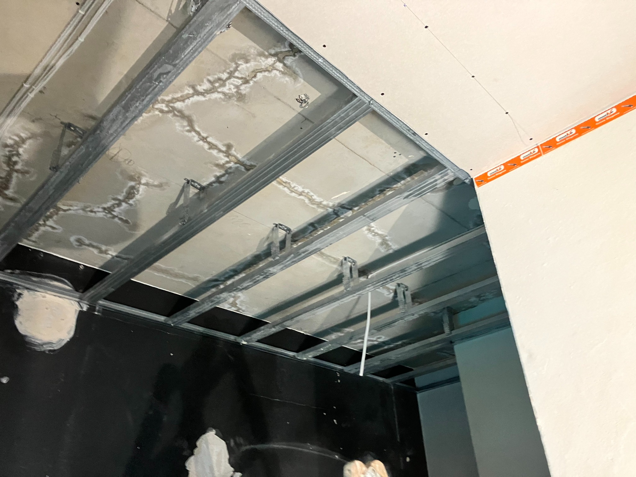 Montaż sufitu podwieszanego z widoczną konstrukcją metalową, częściowo zakrytą płytą gipsowo-kartonową z pomarańczową taśmą uszczelniającą na łączeniu ze ścianą, oraz śladami wilgoci na starym...