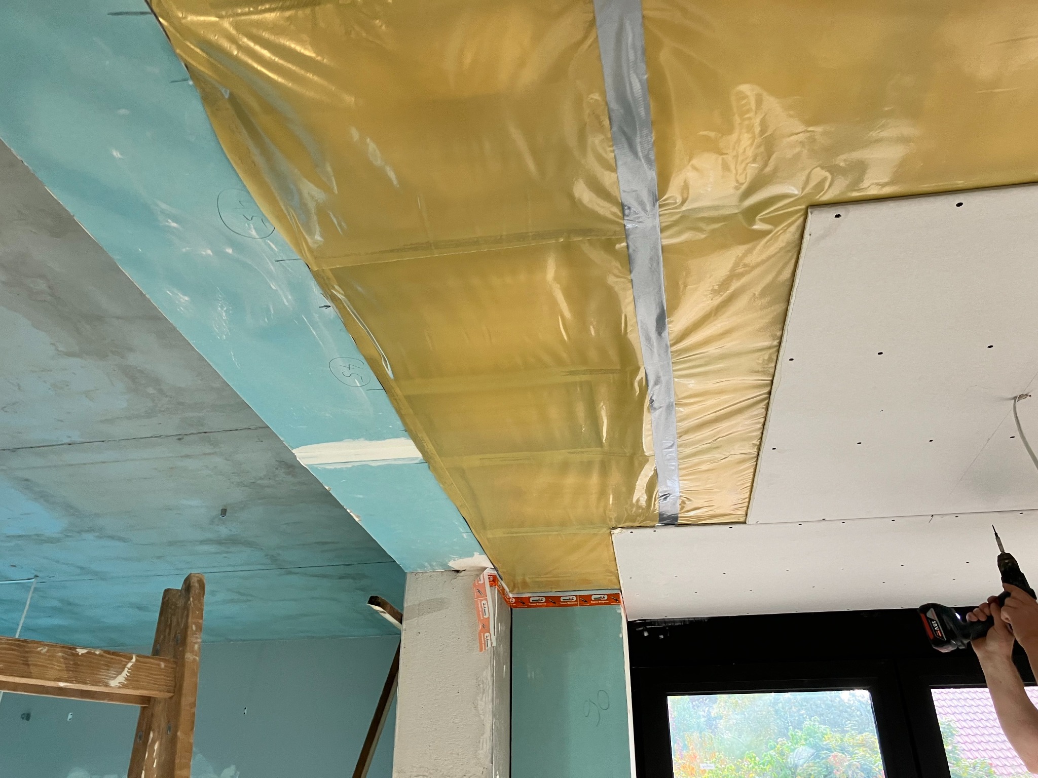 Montaż sufitu podwieszanego: widoczne płyty gipsowo-kartonowe mocowane do folii paroizolacyjnej za pomocą wkrętarki akumulatorowej, fragment drabiny malarskiej w tle.