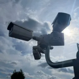 Trzy kamery monitoringu CCTV zamontowane na metalowym wysięgniku, skierowane w różnych kierunkach na tle pochmurnego nieba z przebijającym się słońcem.