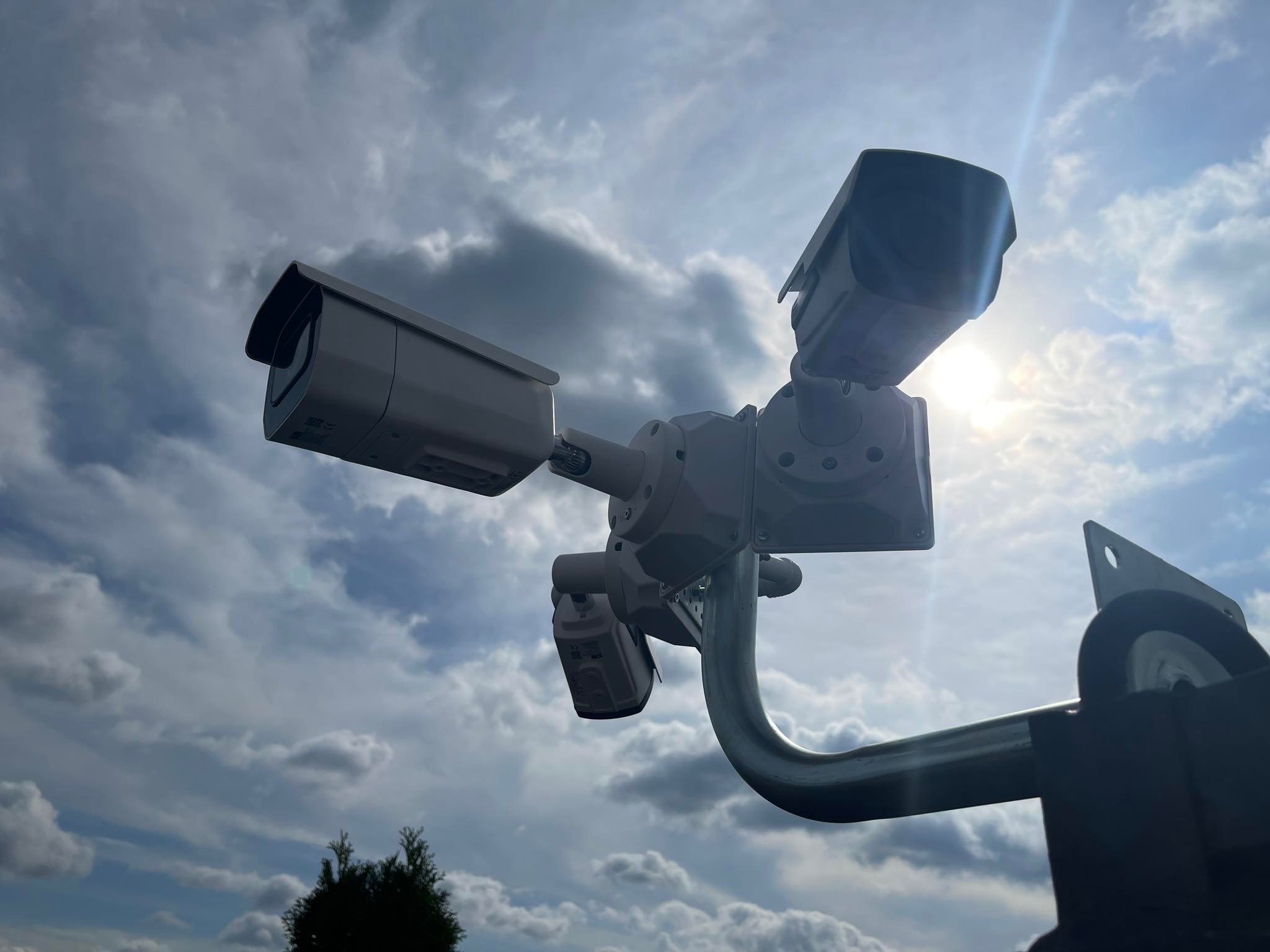 Trzy kamery monitoringu CCTV zamontowane na metalowym wysięgniku, skierowane w różnych kierunkach na tle pochmurnego nieba z przebijającym się słońcem.