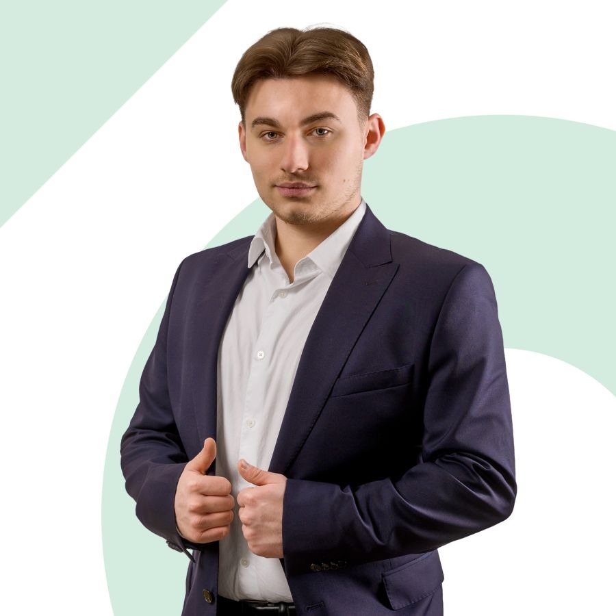 Młody mężczyzna w granatowym garniturze i białej koszuli, z rękami ułożonymi w okolicy zapięcia, na jasnym tle z abstrakcyjnymi, pastelowymi elementami.