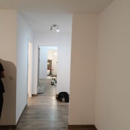 Perfect wykończenia wnętrz Damian Oszczęda - Wykończony korytarz w mieszkaniu po remoncie. Ściany pomalowane na biało, na podłodze panele imitujące drewno. Widok na wyremontowaną łazienkę w głębi.