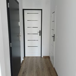 Perfect wykończenia wnętrz Damian Oszczęda - Nowoczesny przedpokój z trzema parami drzwi w kolorach czerni i bieli, drewnopodobna podłoga, minimalistyczne oświetlenie sufitowe.