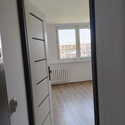 Perfect wykończenia wnętrz Damian Oszczęda - Nowe drzwi wewnętrzne, białe z czarnymi akcentami, zamontowane w odnowionym mieszkaniu. Widok na jasne wnętrze z drewnianą podłogą i oknem.