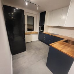 Perfect wykończenia wnętrz Damian Oszczęda - Nowoczesna kuchnia po remoncie: granatowe szafki dolne z drewnianym blatem, białe szafki górne, szara podłoga i czarna lodówka. Minimalistyczne oświetlenie.