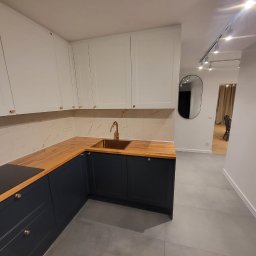 Perfect wykończenia wnętrz Damian Oszczęda - Wykończona kuchnia w bieli i granacie: drewniany blat, złota armatura, marmurowe płytki. Widok na salon z owalnym lustrem. Nowoczesne oświetlenie.