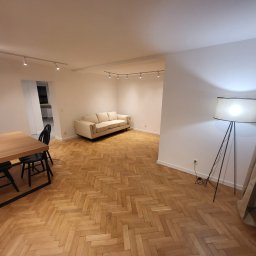 Perfect wykończenia wnętrz Damian Oszczęda - Jasne, przestronne wnętrze po remoncie z parkietem w jodełkę, nowoczesnym oświetleniem, sofą i stołem z krzesłami. Widok na odnowione mieszkanie.