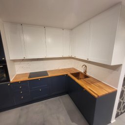 Perfect wykończenia wnętrz Damian Oszczęda - Nowoczesna kuchnia z drewnianym blatem i białymi szafkami górnymi. Widoczny złoty kran i marmurowe wykończenie ściany. Ciemne szafki dolne z złotymi uchwytami.