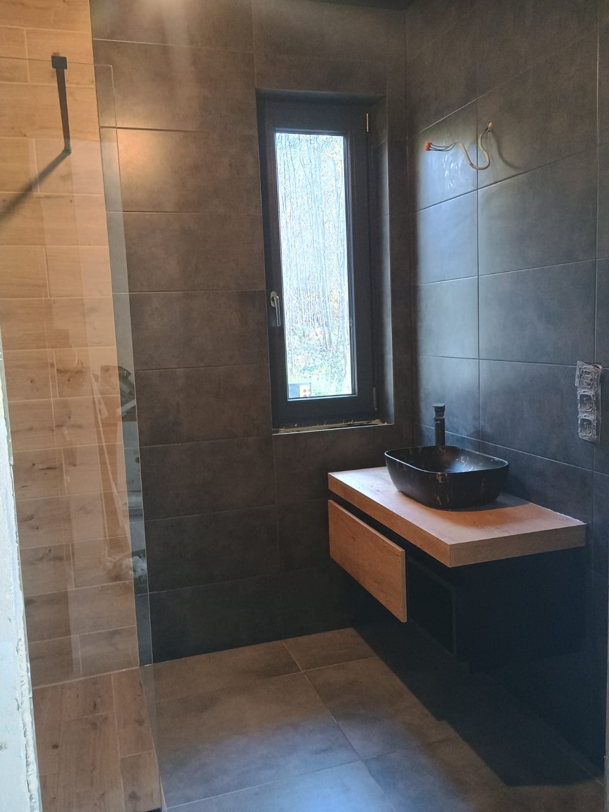 Nowoczesna łazienka z ciemnymi płytkami, drewnianą szafką pod umywalką i oknem z widokiem na drzewa, w trakcie wykańczania.