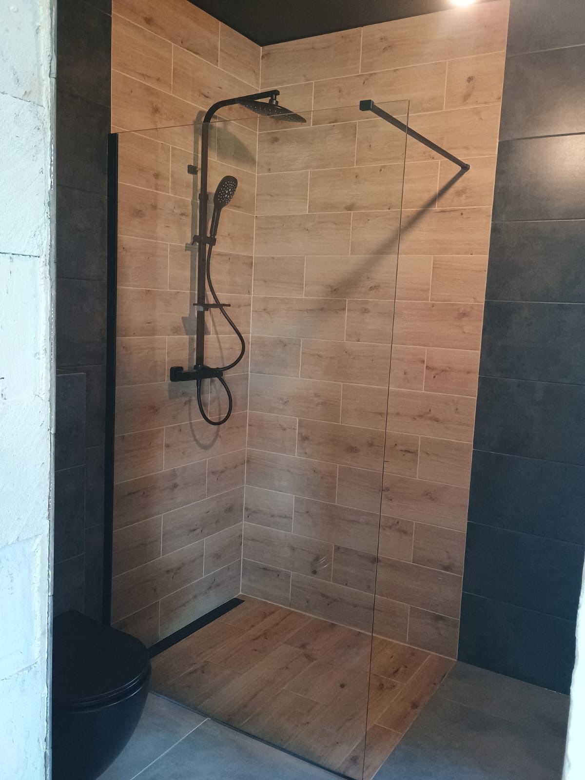 Nowoczesna kabina prysznicowa walk-in z czarną armaturą na tle płytek imitujących drewno i szarych płytek. Minimalistyczny design, widoczny odpływ liniowy.