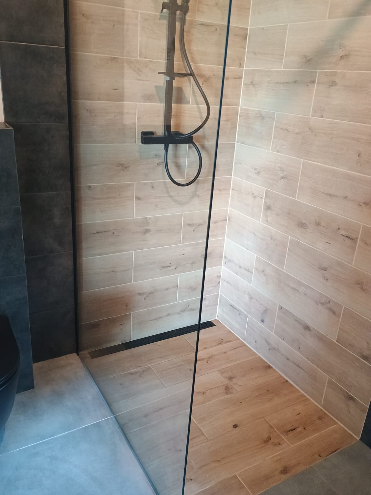 Kabina prysznicowa walk-in z drewnopodobnymi płytkami, czarną armaturą i odpływem liniowym. Minimalistyczny design, jasne płytki na ścianach i podłodze.