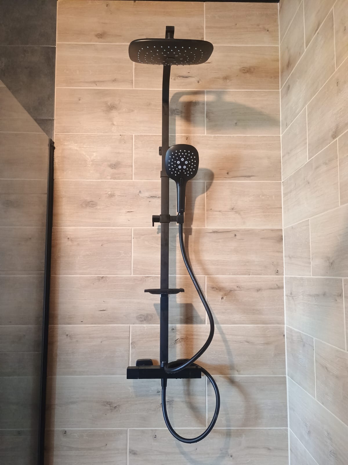 Nowoczesny czarny panel prysznicowy z deszczownicą i słuchawką, zamontowany na ścianie wyłożonej płytkami imitującymi drewno w wykończonej łazience.