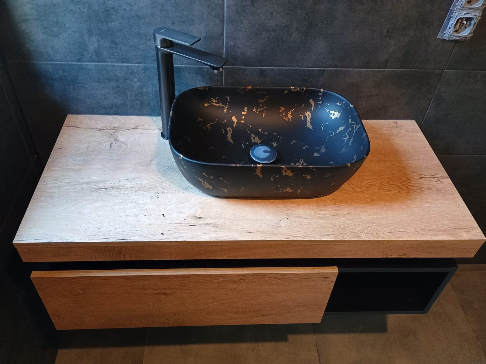 Czarna, nablatowa umywalka z armaturą na drewnianej szafce w łazience z szarymi płytkami. Umywalka zdobiona złotymi wzorami.