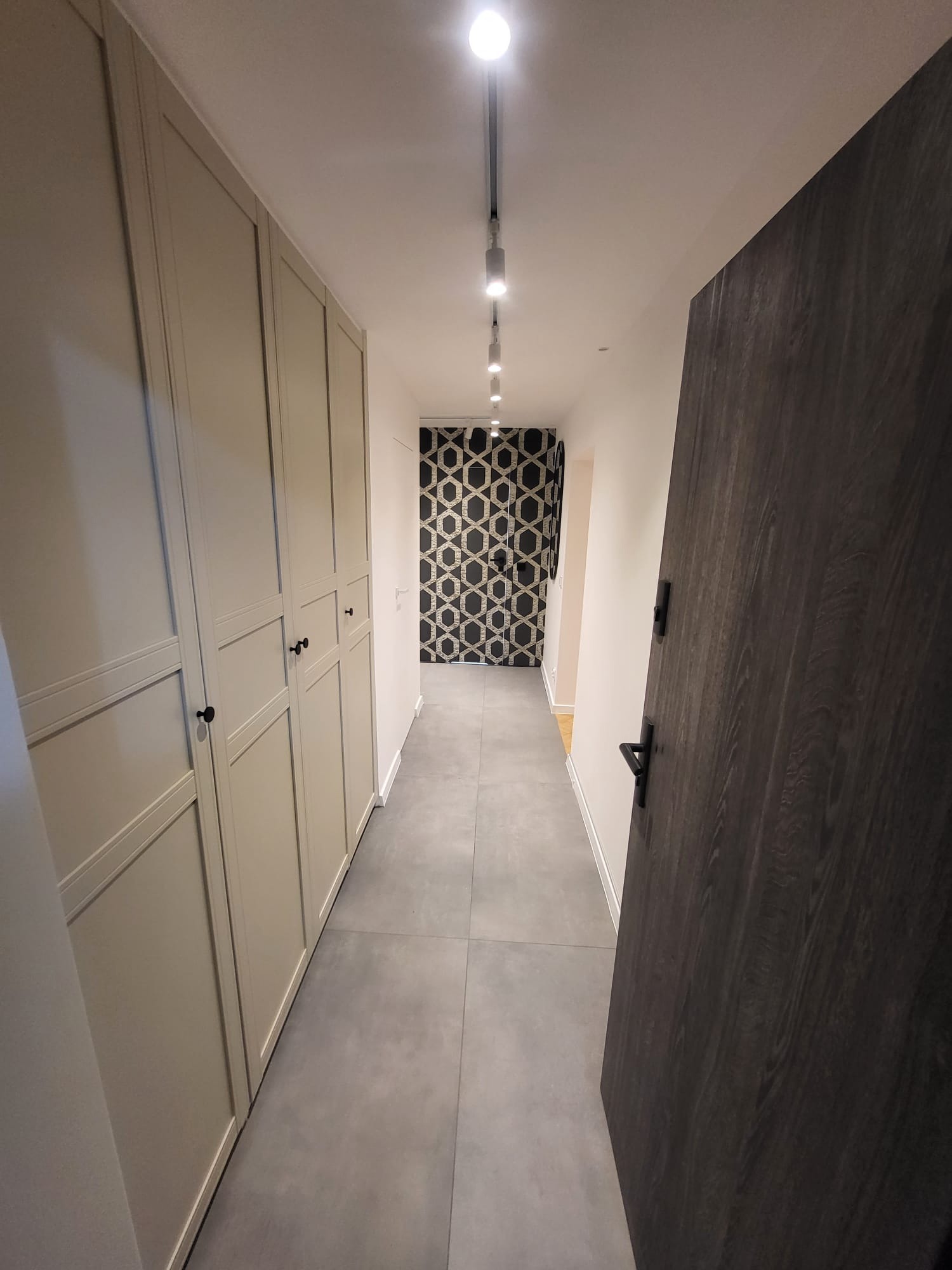 Korytarz po remoncie: szara podłoga, zabudowa szafy w kolorze beżowym, ciemne drzwi z klamką, oświetlenie sufitowe i wzorzysta tapeta w głębi.