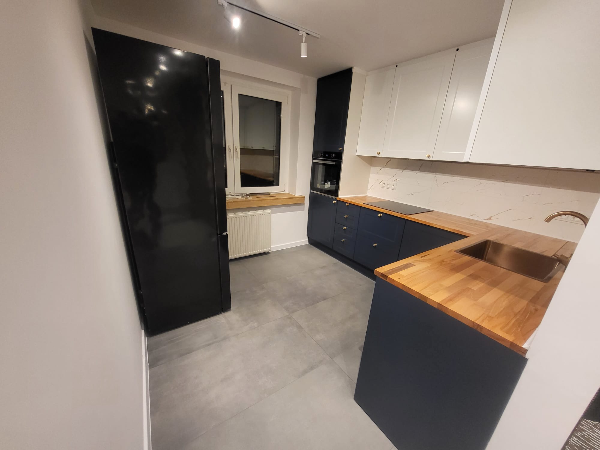 Nowoczesna kuchnia po remoncie: granatowe szafki dolne z drewnianym blatem, białe szafki górne, szara podłoga i czarna lodówka. Minimalistyczne oświetlenie.