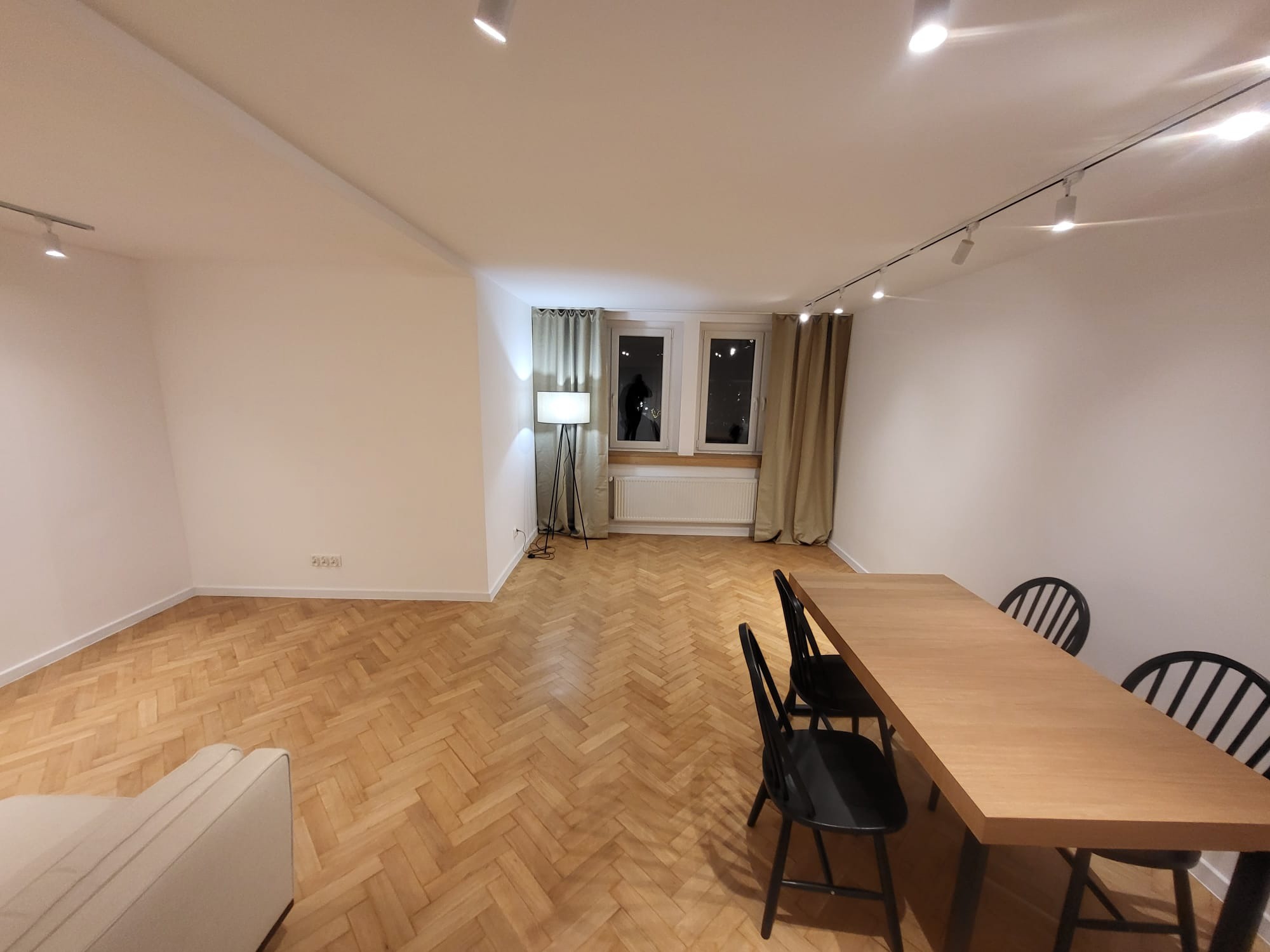 Jasne, przestronne wnętrze po remoncie: parkiet w jodełkę, białe ściany, nowoczesne oświetlenie szynowe, drewniany stół z krzesłami i sofa w rogu.