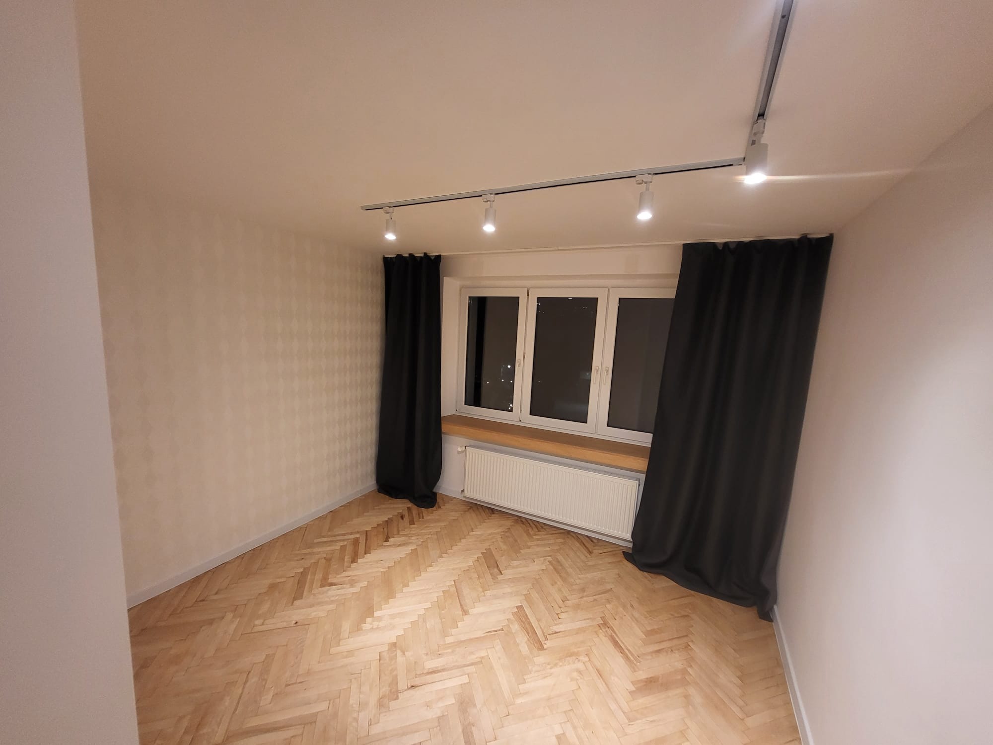 Pokój po remoncie: parkiet w jodełkę, okno z parapetem, grzejnik, szyny z oświetleniem punktowym i zasłony. Ściany z delikatnym wzorem. Widok na wnętrze.